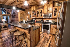 A rustic kitchen with wooden cabinetry, a central island with stools, stainless steel appliances, and a large refrigerator. - Stairway to Heaven 7BR Luxury Mountain Views (Broken Bow)