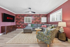 A living room with a sectional sofa, a patterned armchair, a TV mounted on the wall, and a ceiling fan. - Sleeps 16 Golf Simulator Pickleball Pool Spa (Bradenton)