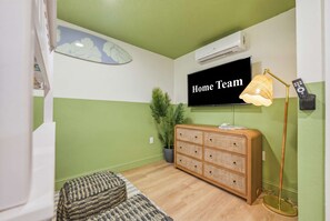 Een kamer met een televisie waarop 'Home Team' te zien is, een rieten salontafel, een vloerlamp en een potplant.