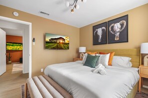 A hotel room with a large bed, a painting of a sunset, two framed animal skulls, and a door leading to another room. - Poolside Pickleball Heated Pool Sleeps 20 (San Antonio)