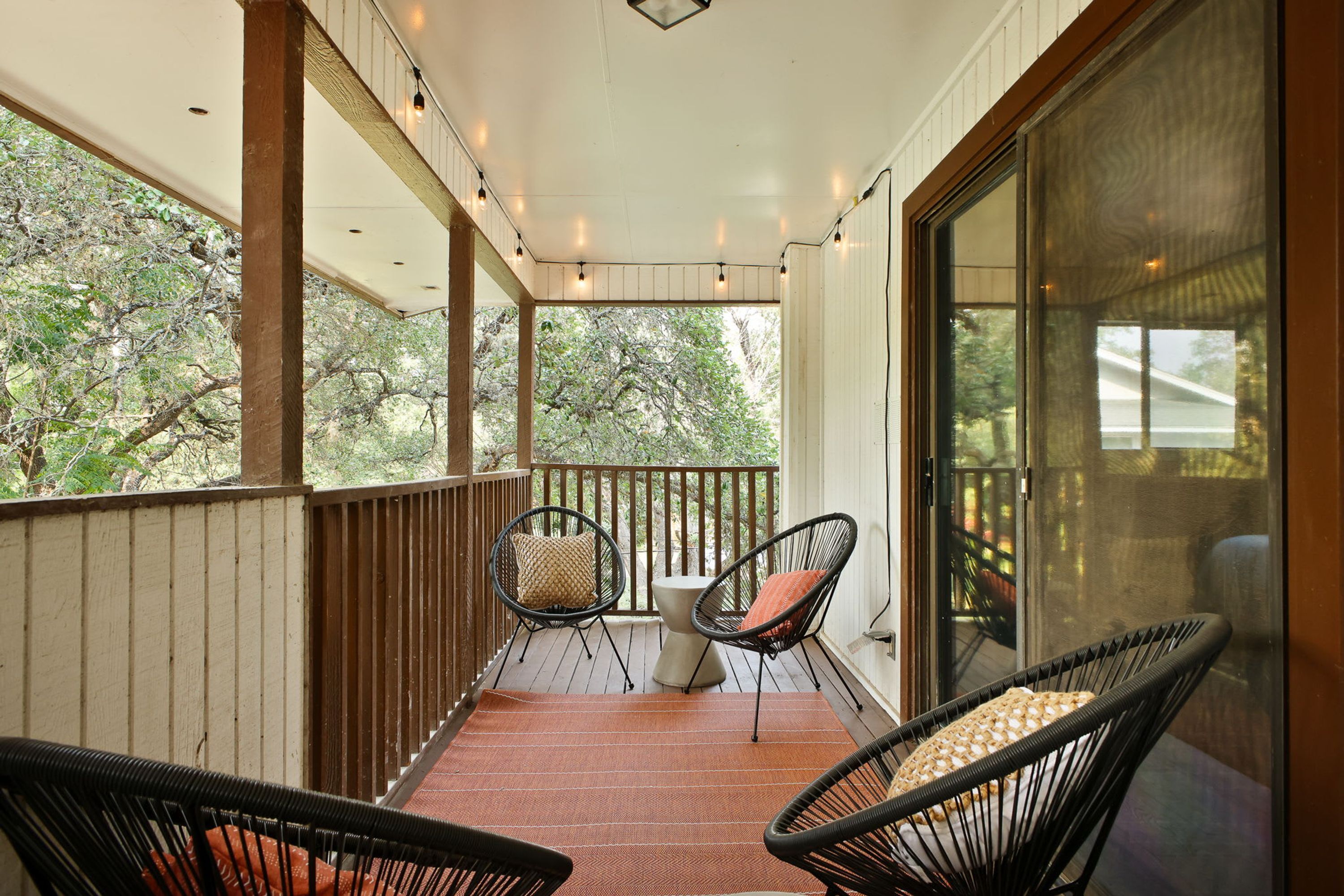A covered porch with two chairs and a table, wooden railings, and a view of trees.