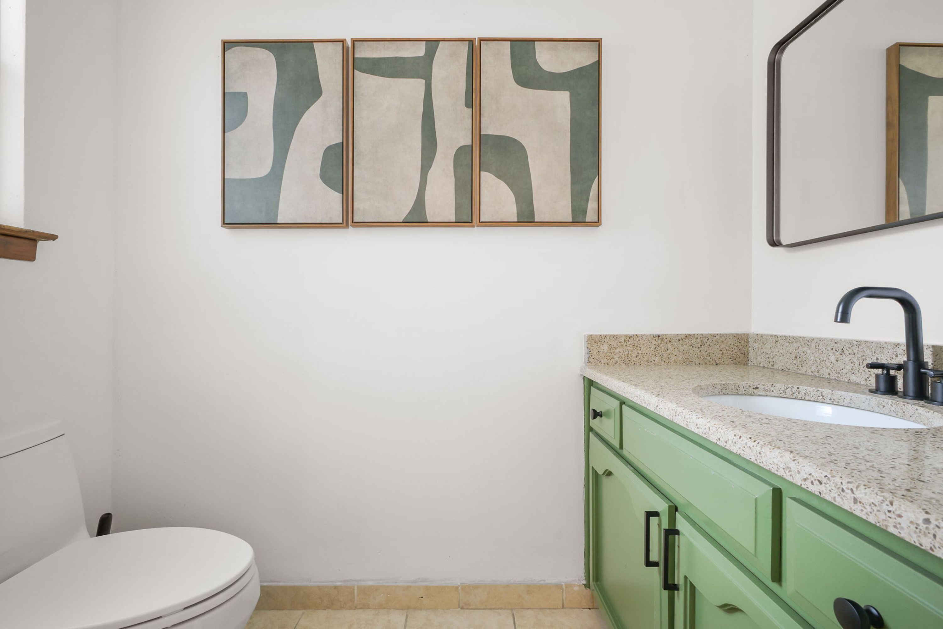 A bathroom with a toilet, a sink, and a mirror. There are three framed abstract paintings on the wall.