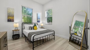 A bedroom with a bed, two bedside tables, a mirror, and artwork on the walls. - Pineapple Paradise - 13 min to Beach - Fire Pit (Sarasota)