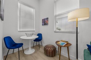 Ein Zimmer mit zwei weißen, runden Tischen, einer mit einem Drahtkorb und einer mit einem Buch. Zwei blaue Stühle, ein brauner Hocker und eine Stehlampe sind ebenfalls vorhanden.