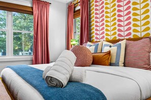 A bedroom with a large bed, multiple pillows, a blue blanket, and a window with curtains. - Outdoor Games Hot Tub Firepit Grill Sauna (Asheville)