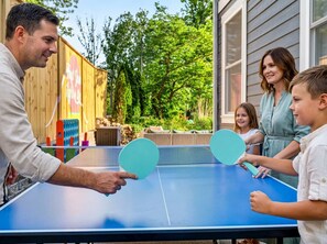 A family playing ping pong outdoors with a wooden fence and greenery in the background. - Outdoor Games Hot Tub Firepit Grill Sauna (Asheville)