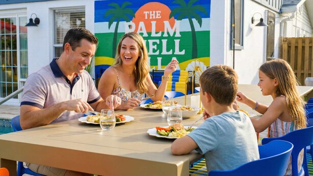 Een gezin dineert aan een buitentafel met uitzicht op een gebouw, waarop een bord staat met de tekst 'THE PALM WELL'.