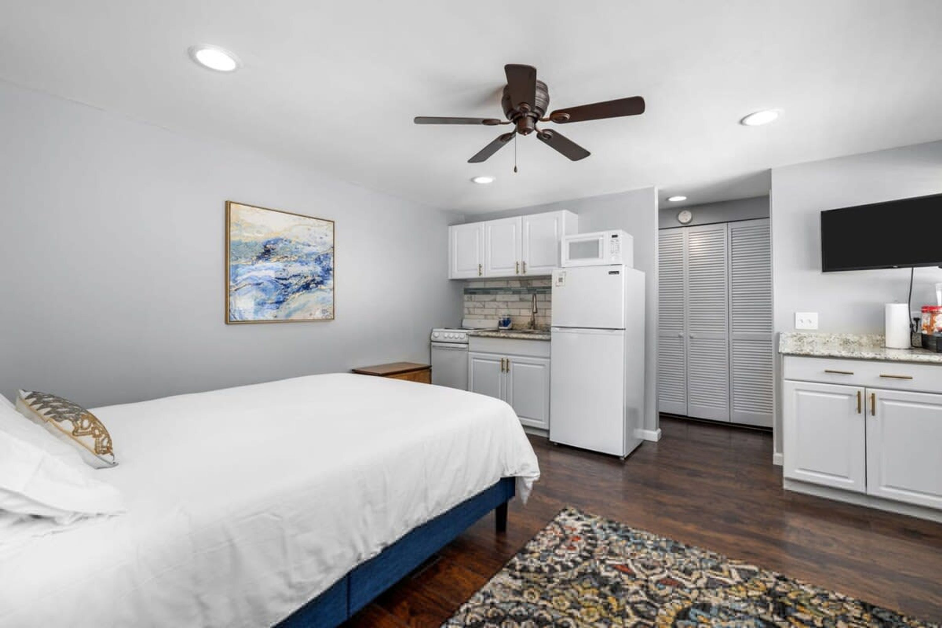 A bedroom with a bed, a ceiling fan, a kitchen with cabinets, a microwave, and a TV.