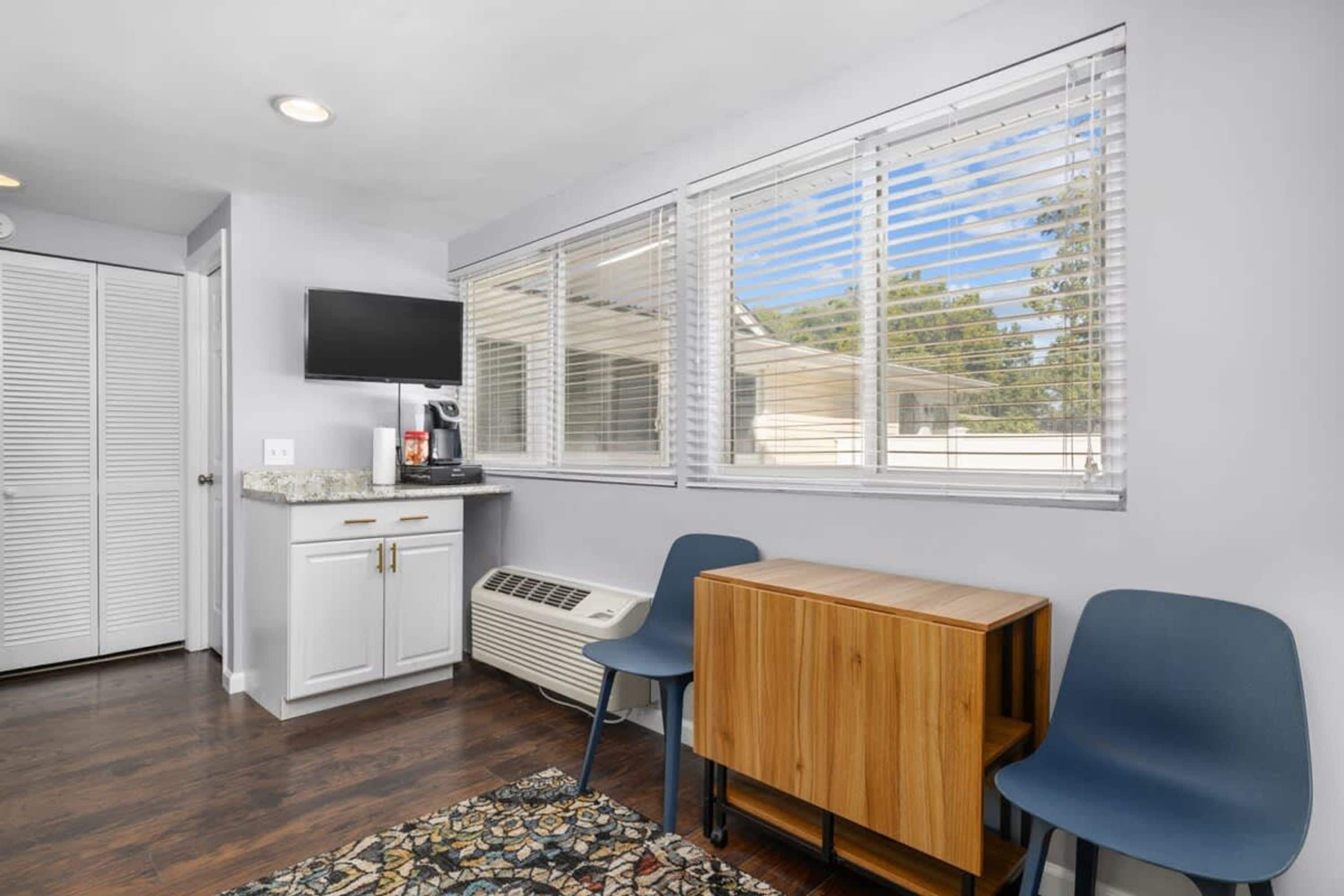 A compact room with a kitchenette, a television mounted on the wall, a window with blinds, and a patterned rug on the floor.