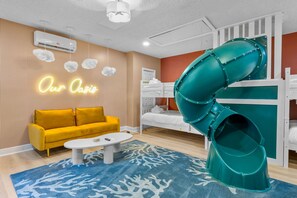 A modern hotel room with a yellow sofa, a white coffee table, a blue rug, a green slide, and a neon sign. - Sleeps 10 Pool Slide Pickleball Fire Pit (Seminole)