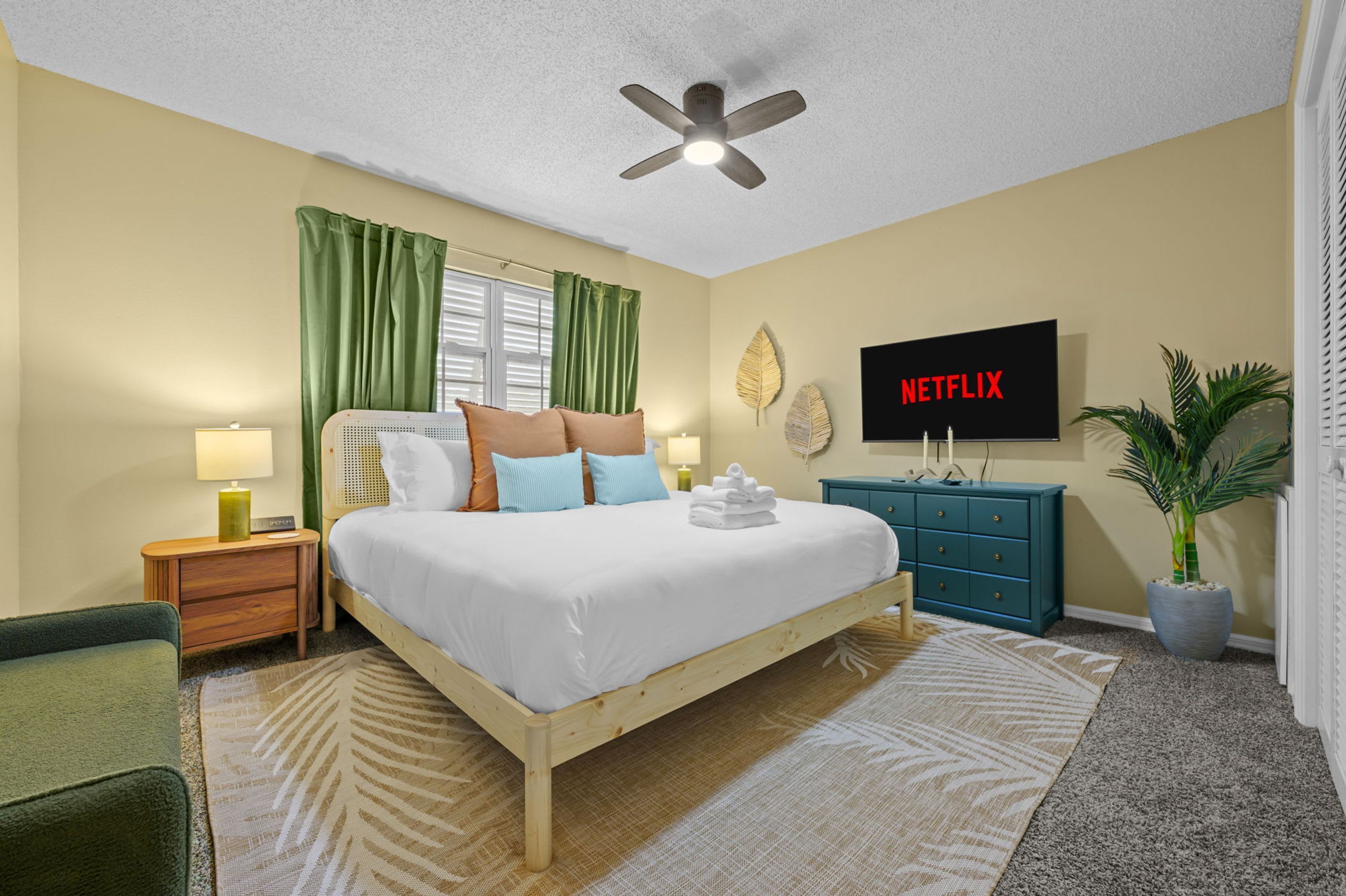 Habitación de hotel con cama, televisor, ventilador de techo, mesita de noche, cómoda, una silla verde, planta en maceta y ventana con cortinas.