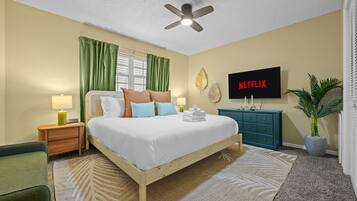 Habitación de hotel con cama, televisor, ventilador de techo, mesita de noche, cómoda, una silla verde, planta en maceta y ventana con cortinas.