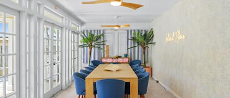 A dining area with a wooden table and blue chairs, a ceiling fan, and a neon sign on the wall.