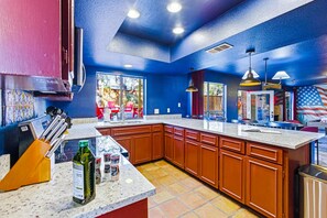 A kitchen with blue walls, wooden cabinets, a granite countertop, and a window overlooking a backyard. - Cowboy Ridge 4BR Spacious Retreat Sleeps 13 (San Antonio)