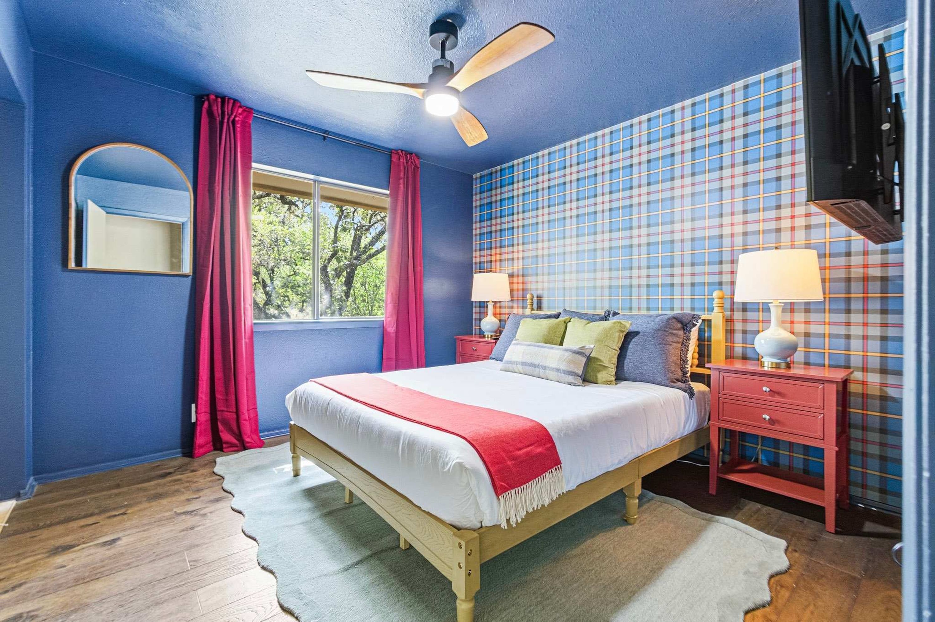 Une chambre à coucher avec un lit, un ventilateur de plafond, une fenêtre avec des rideaux, une table de chevet avec une lampe et un mur orné d’un motif à carreaux.