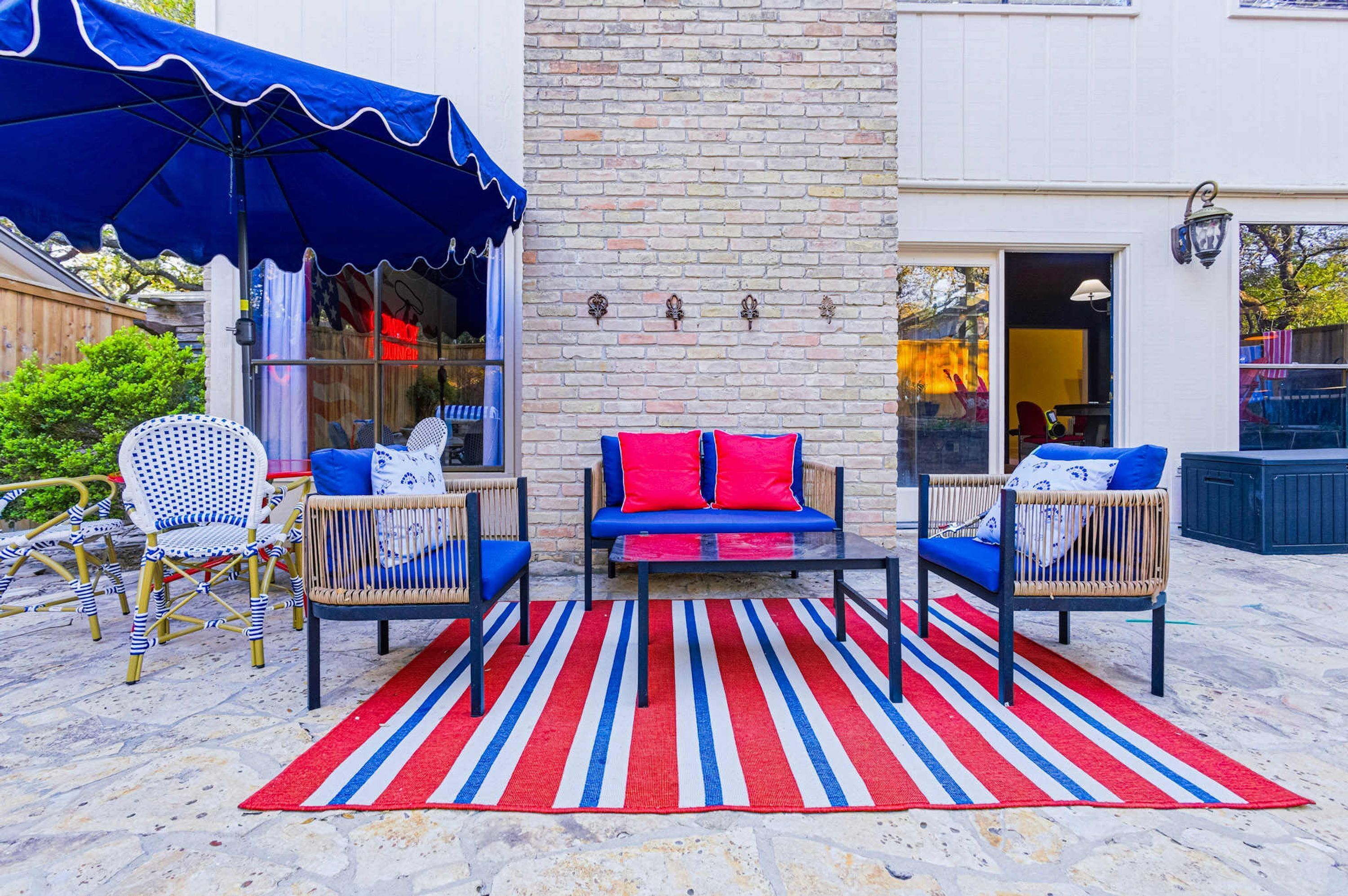 An outdoor seating area with a red and blue striped rug, wicker chairs, and a blue umbrella.
