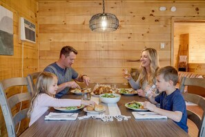 A family dining in a wooden cabin with a table set for a meal, a hanging light fixture, and a window with a vent.