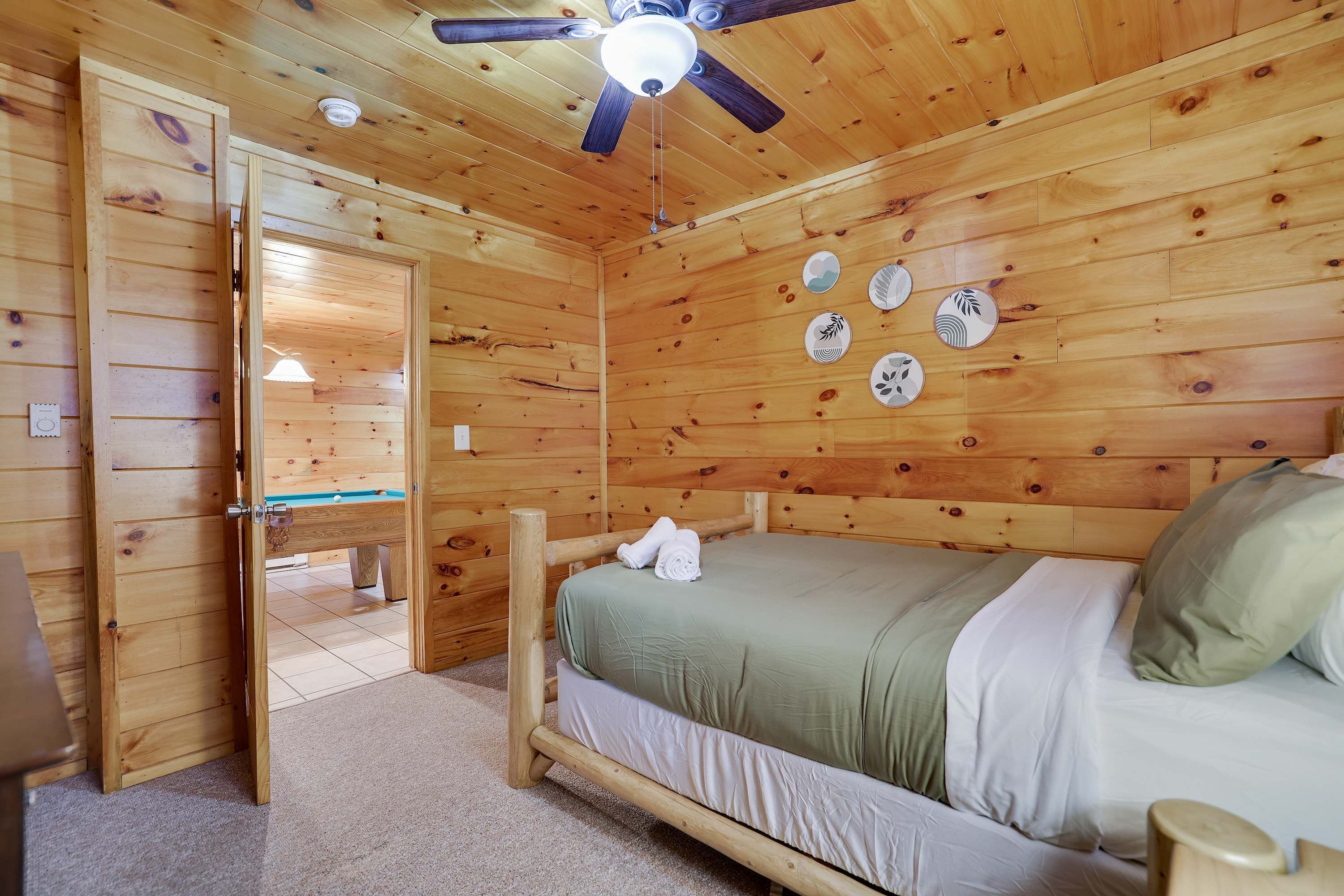 A wooden room with a bed, a ceiling fan, and a door leading to another room.