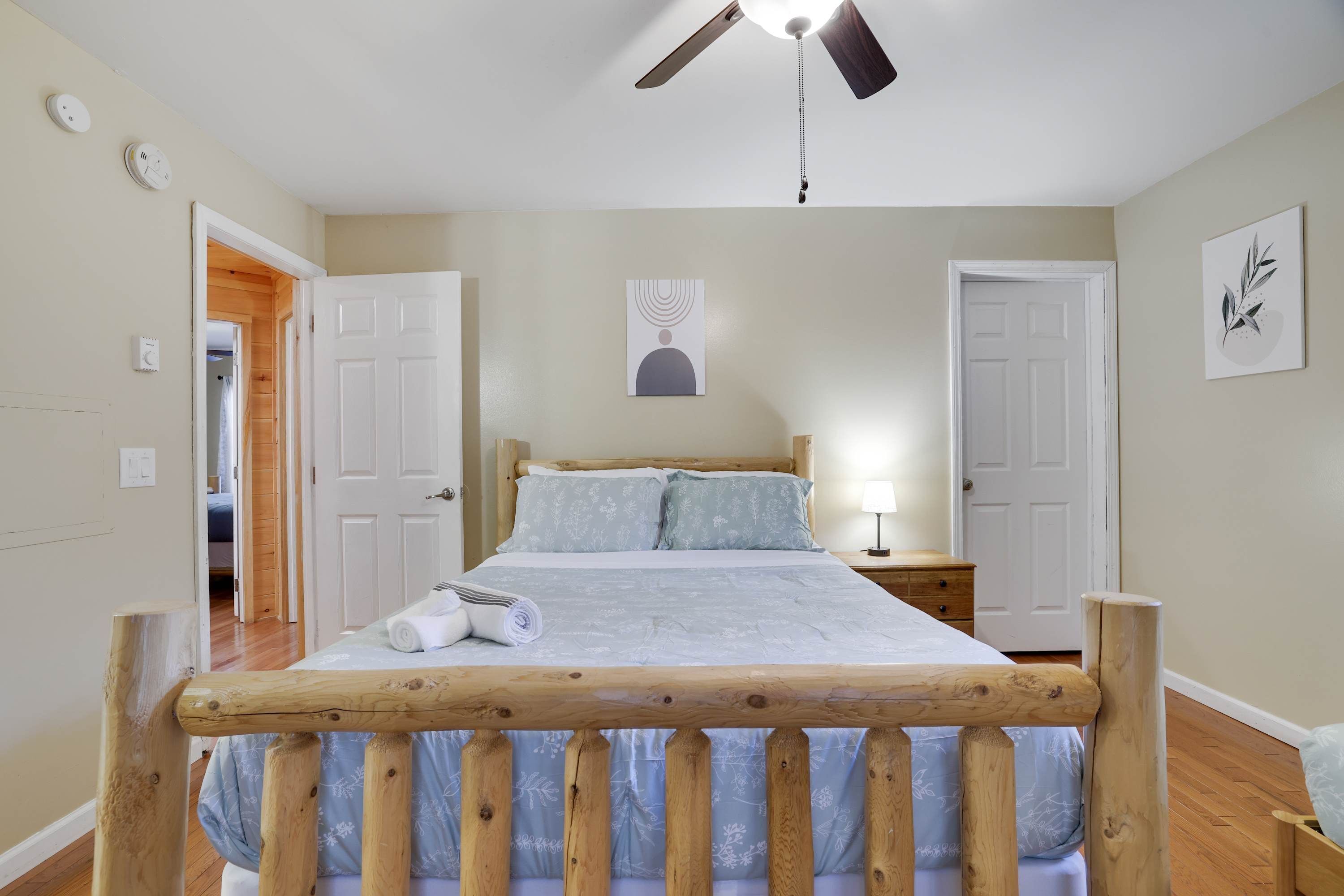 Un dormitorio con estructura de cama de madera, ventilador de techo, mesita de noche con lámpara y puerta que da a otra habitación.