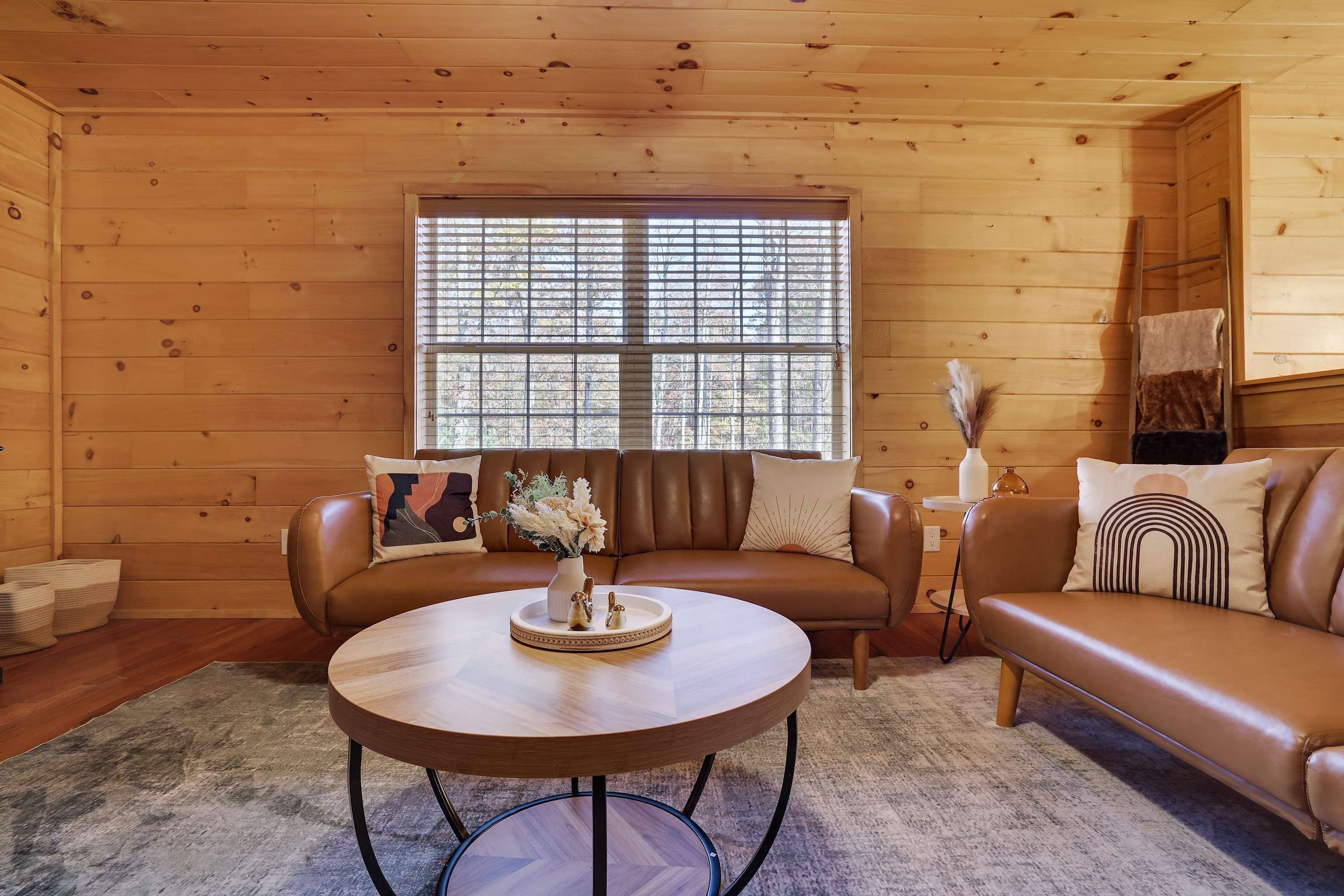 A cozy cabin interior with a brown leather sofa, a round wooden table, and a large window with blinds.