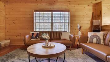 A cozy cabin interior with a brown leather sofa, a round wooden table, and a large window with blinds.