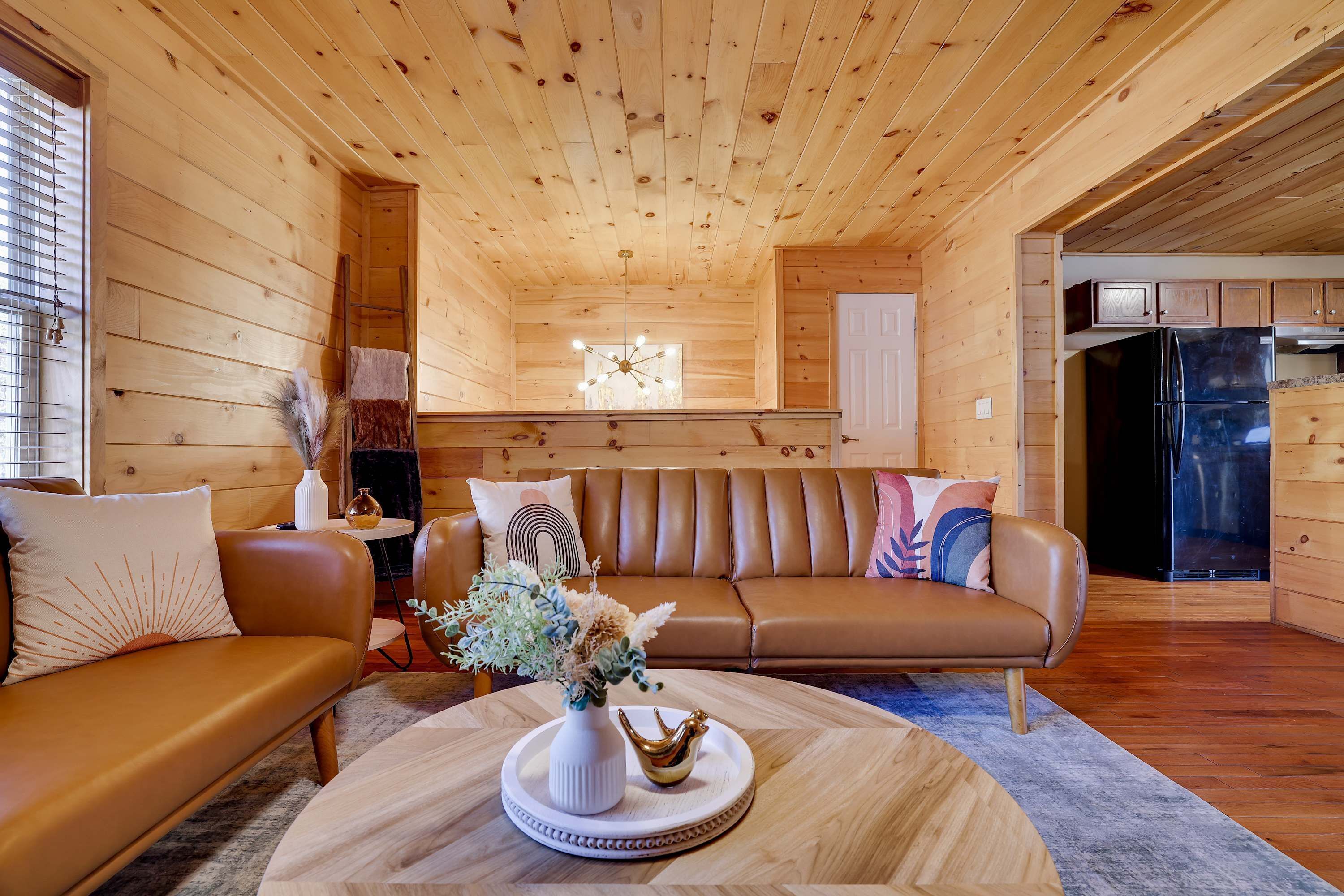 A cozy cabin interior with a wooden ceiling, a brown leather sofa, a coffee table with decorative items, and a kitchen area visible through an open doorway.