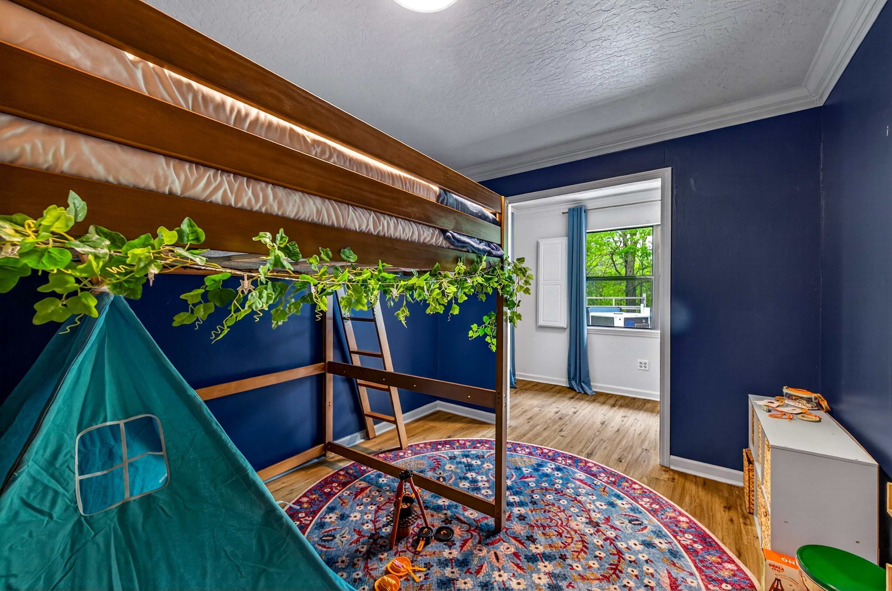 Een kamer met een blauwe wand, een houten hoogslaper met een hangende plant en een groene bladeren, een kleurrijk vloerkleed en een trap.