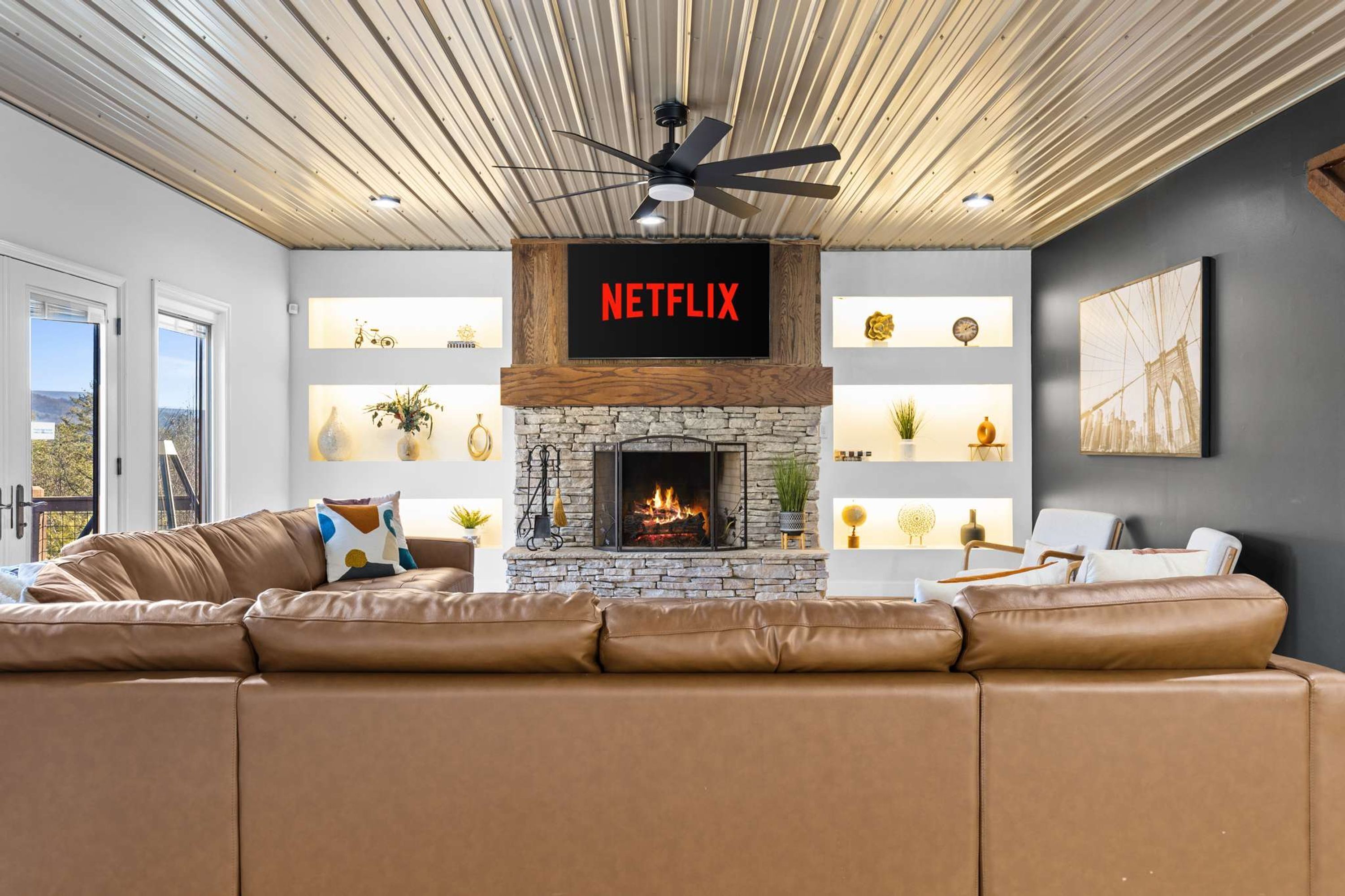 A living room with a stone fireplace, a Netflix sign, a ceiling fan, and a large window.