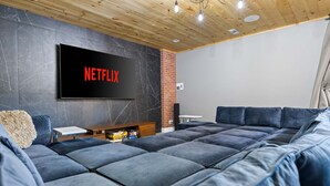 Uma sala de estar moderna com um amplo sofá modular, uma TV de tela plana fixada em uma parede de mármore escuro e teto de madeira com iluminação embutida.