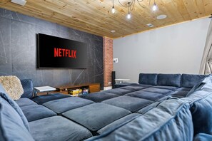 A modern living room with a large sectional sofa, a flat-screen TV mounted on a dark marble wall, and a wooden ceiling with recessed lighting. - Mtn Views Theater Hot Tub 5 King Suites (Blue Ridge)
