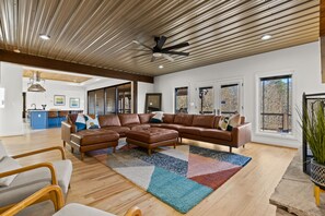 A spacious living area with a large sectional sofa, a ceiling fan, and a view of the outdoors through large windows. - Mtn Views Theater Hot Tub 5 King Suites (Blue Ridge)