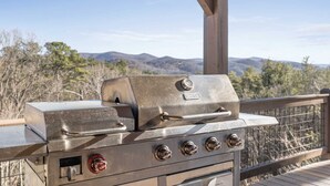 Uma churrasqueira de aço inoxidável para áreas externas, com quatro queimadores e um queimador lateral, instalada em um deck de madeira com vista para montanhas ao fundo.