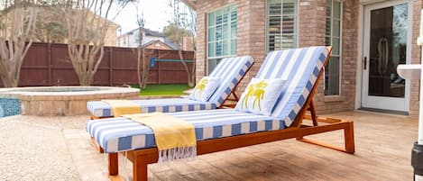 A deck with two lounge chairs, a small pool, and a brick house in the background.