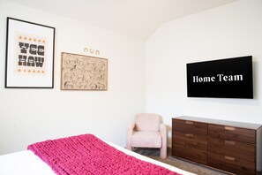 A bedroom with a bed, a pink chair, a wooden dresser, and a TV on the wall. - Upscale Escape Pool Hot Tub Game Room Firepit (San Antonio)