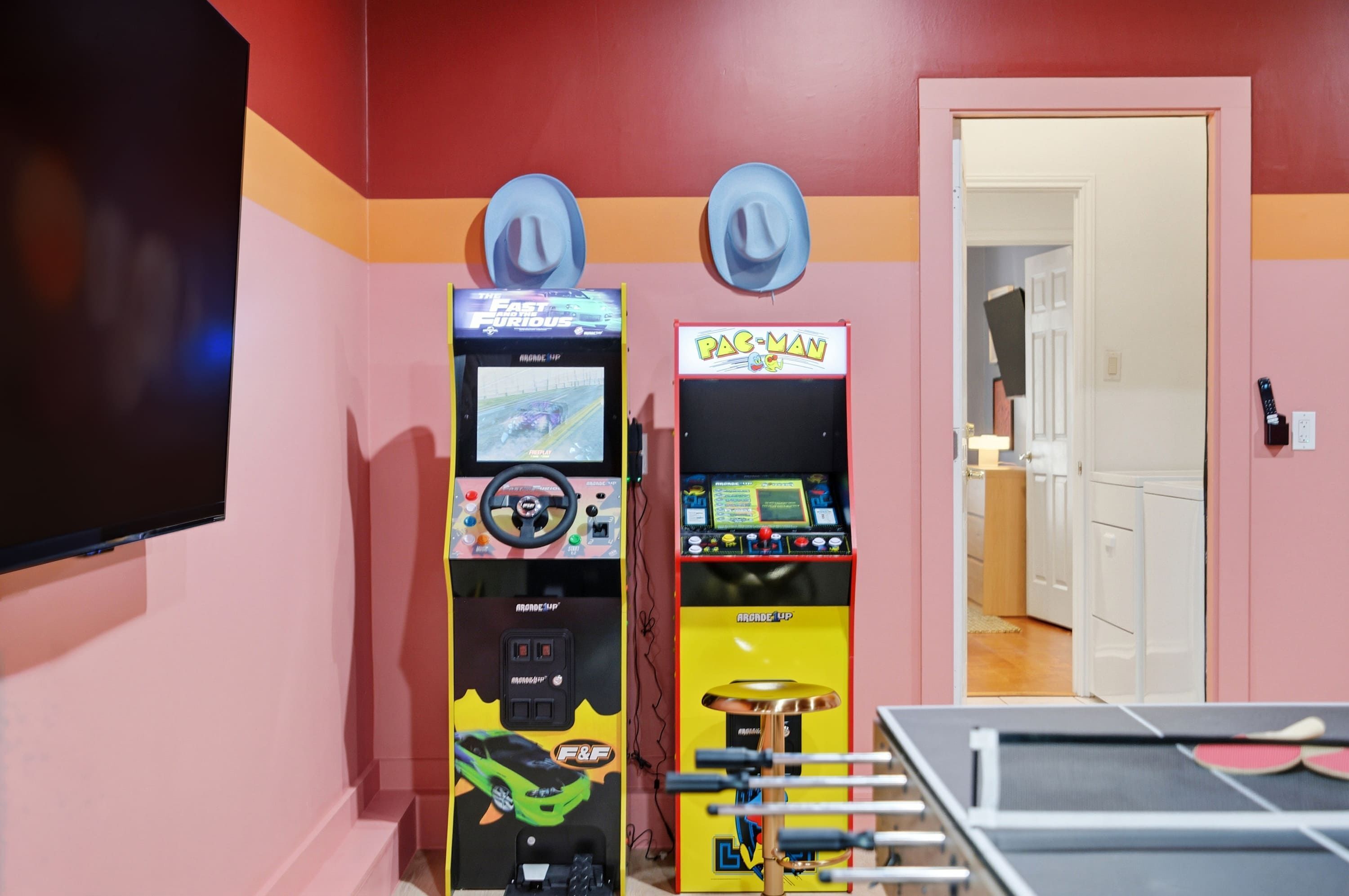 Two arcade machines, one for 'The Fast and the Furious' and another for 'Pac-Man', in a room with a pink wall and a door leading to another room.