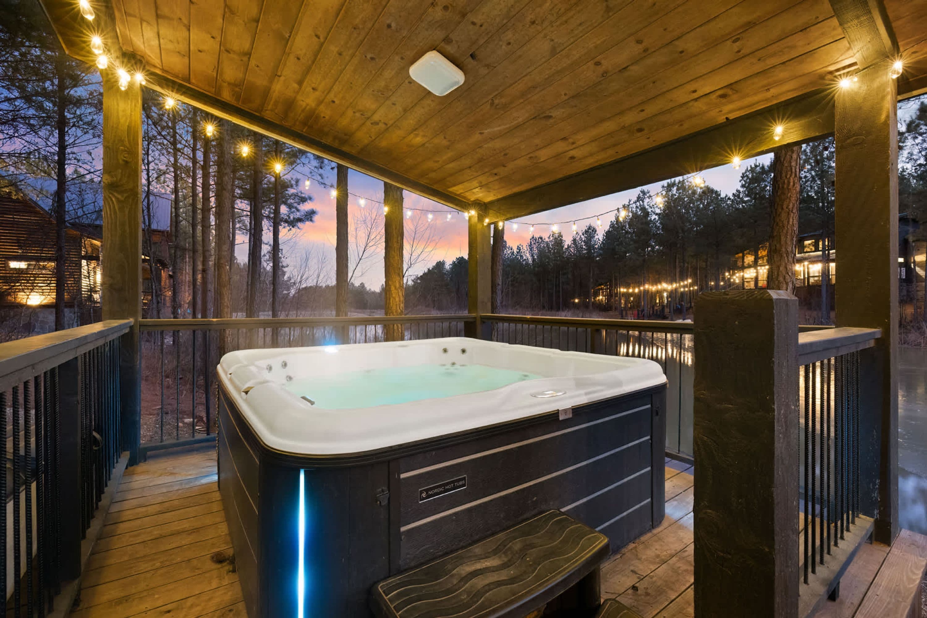 A hot tub on a wooden deck with string lights, surrounded by trees and a cabin in the background.