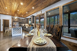 A spacious dining area with a long table set for a meal, large windows offering a view of the outdoors, and a kitchen area with modern appliances. - The Waterside Retreat Hot Tub Kayaks EV Charger (Broken Bow)