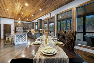 A spacious dining area with a long table set for a meal, large windows offering a view of the outdoors, and a kitchen area with modern appliances.