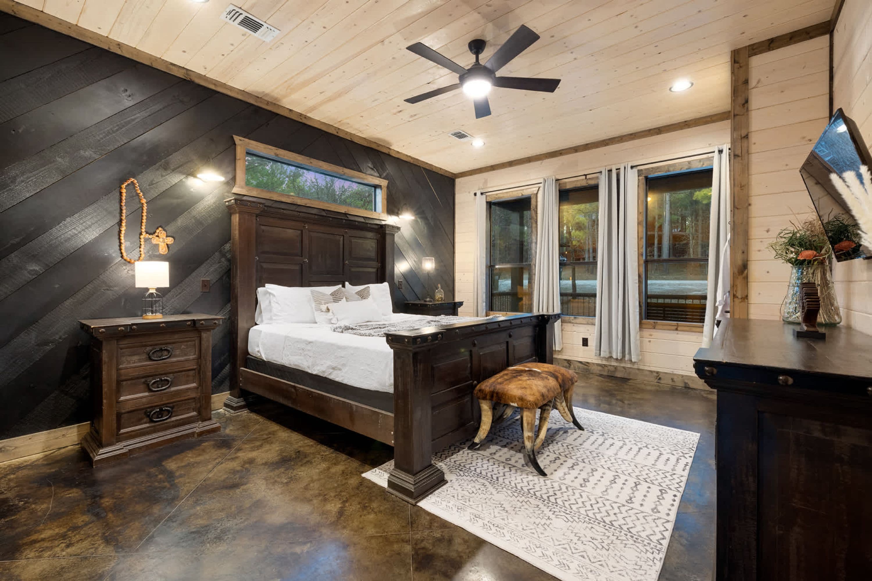 A bedroom with a wooden ceiling, a bed with white linens, a nightstand, a lamp, a window with curtains, and a view of the outdoors.