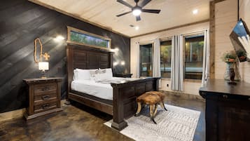 A bedroom with a wooden ceiling, a bed with white linens, a nightstand, a lamp, a window with curtains, and a view of the outdoors.