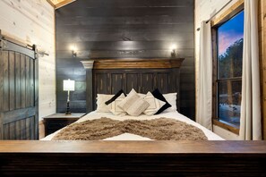 A bedroom with a wooden headboard, a bed with multiple pillows, a nightstand with a lamp, and a window with a view of trees. - The Waterside Retreat Hot Tub Kayaks EV Charger (Broken Bow)
