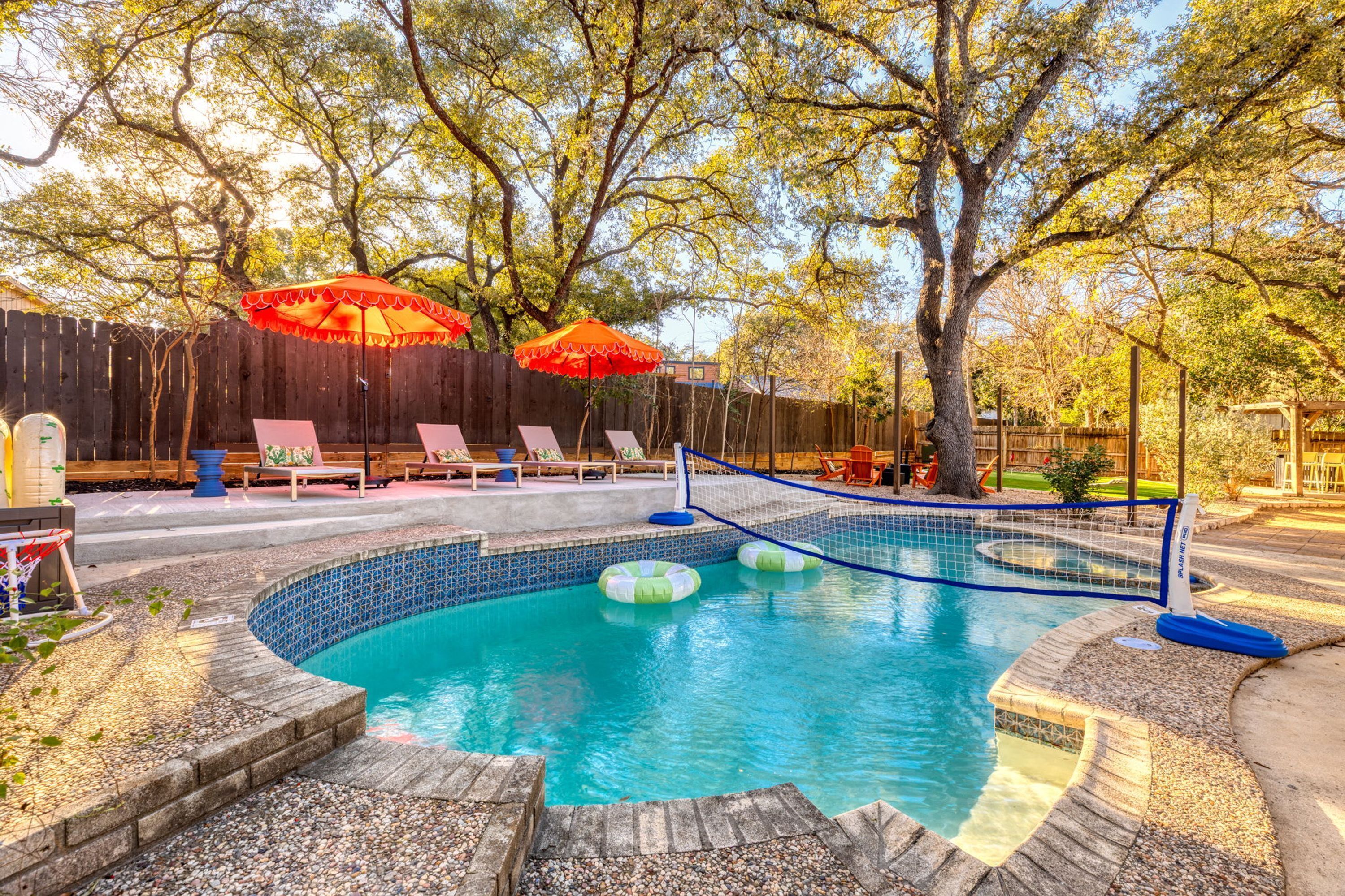 A swimming pool with a tiled edge, surrounded by a pebble deck, and shaded by two red umbrellas.