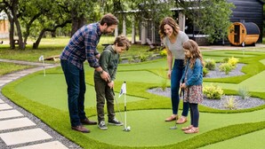 Uma família jogando minigolfe num jardim bem cuidado, com um green de putting, um barril de madeira e um pequeno lago.