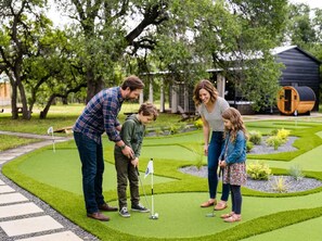 Eine Familie spielt Minigolf in einem gepflegten Garten mit Putting Green, einem Holzbottich und einem kleinen Teich.