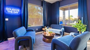 A modern lounge area with dark blue walls, a neon sign reading 'SQUEEZE THE DAY', and a large window offering a view of the outdoors. - Suncrest Retreat - Pool Pickleball Theater (Scottsdale)