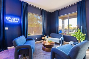A modern lounge area with dark blue walls, a neon sign reading 'SQUEEZE THE DAY', and a large window offering a view of the outdoors. - Suncrest Retreat - Pool Pickleball Theater (Scottsdale)