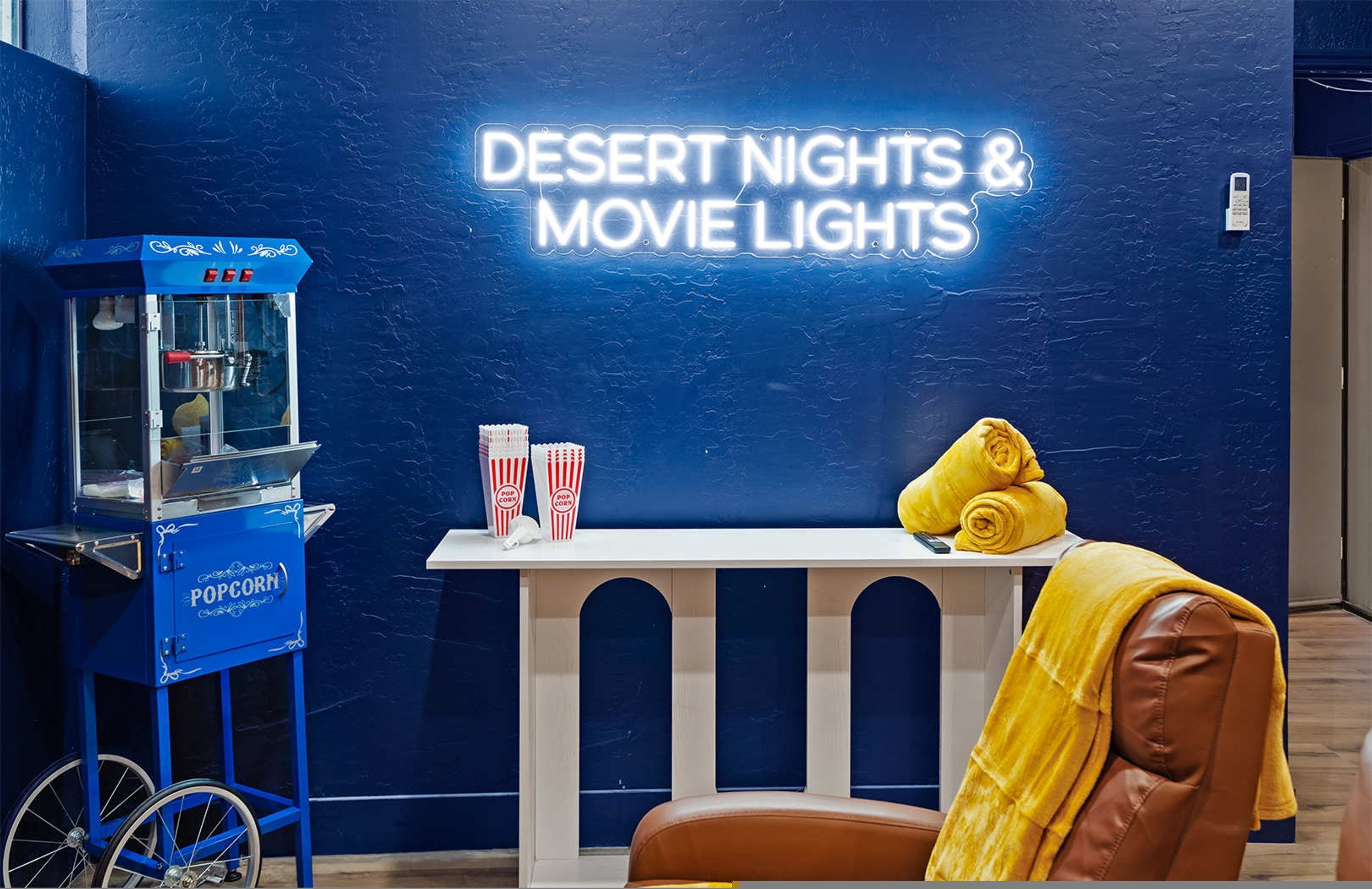 A blue popcorn machine labeled 'POP CORN' and a neon sign reading 'DESERT NIGHTS & MOVIE LIGHTS' on a blue wall.