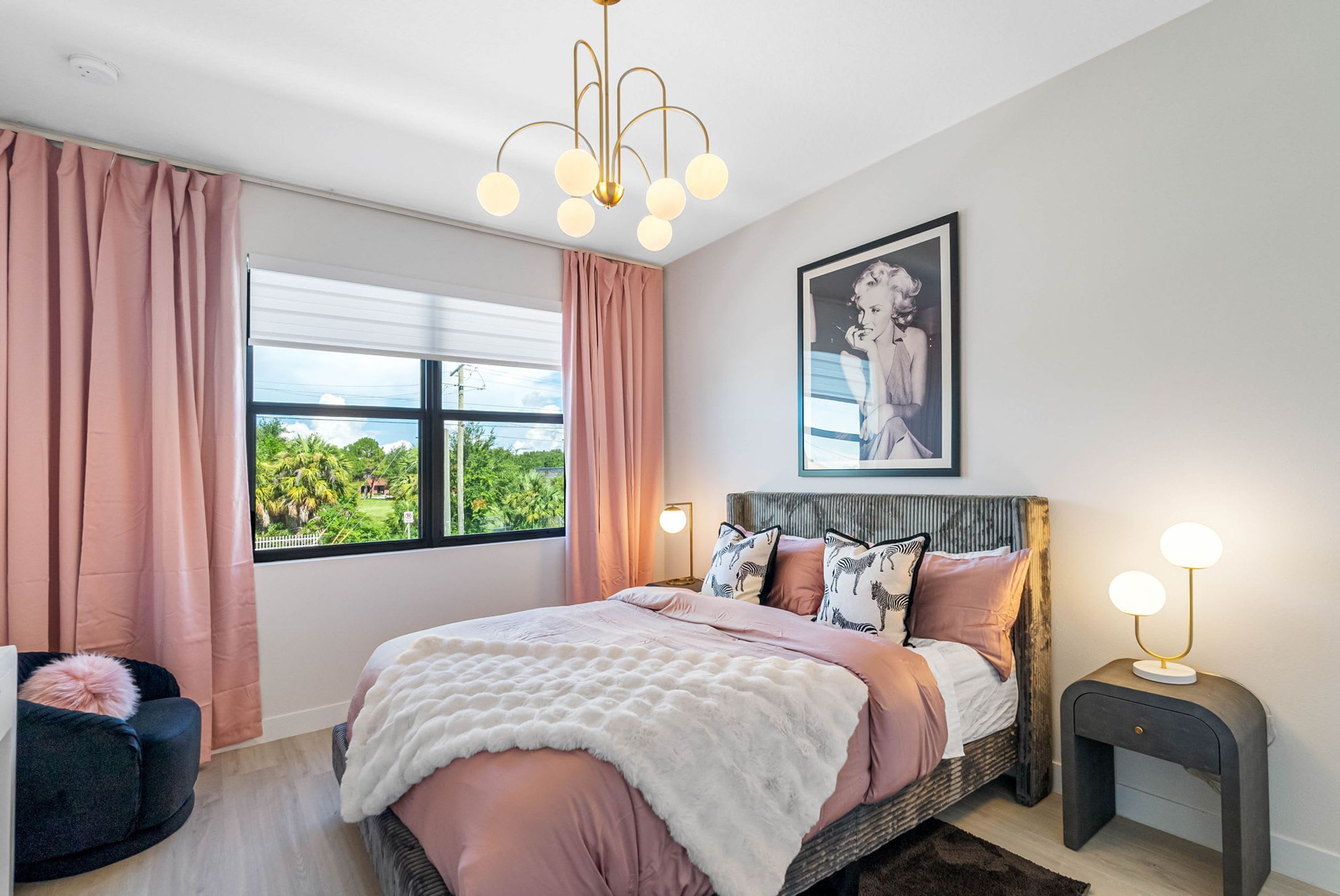 Quarto com cama, janela com cortinas, candelabro, quadro na parede e mesa de cabeceira com abajur.
