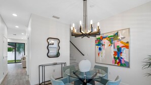 A modern dining area with a glass table and blue chairs, a chandelier, a large abstract painting, and a staircase. - Brand New Tampa Townhouse Prime Location (Tampa)