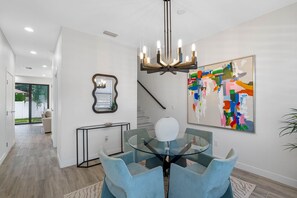 A modern dining area with a glass table and blue chairs, a chandelier, a large abstract painting, and a staircase. - Brand New Tampa Townhouse Prime Location (Tampa)